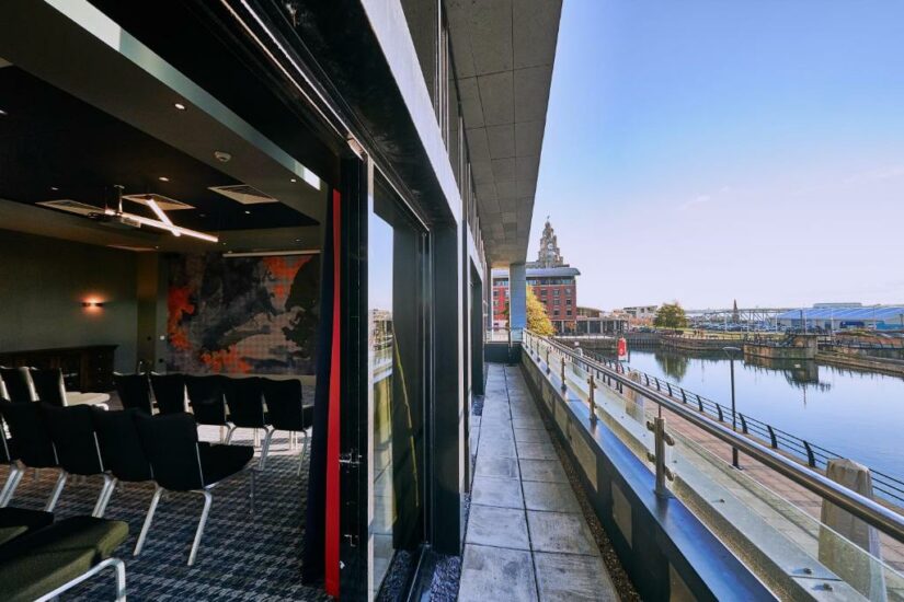 Uma sala de reuniões com cadeiras fica de frente para portas de vidro abertas que levam a uma varanda com vista para a orla e a paisagem urbana.