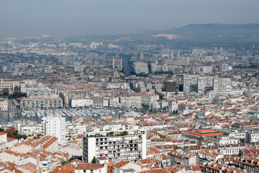 onde ficar em Marselha em Notre-Dame du Mont e Cours Julien Vista aérea de do bairro Notre-Dame du Mont e Cours Julien em Marseille, mostrando edifícios com telhados vermelhos, uma marina e colinas distantes sob um céu nebuloso.