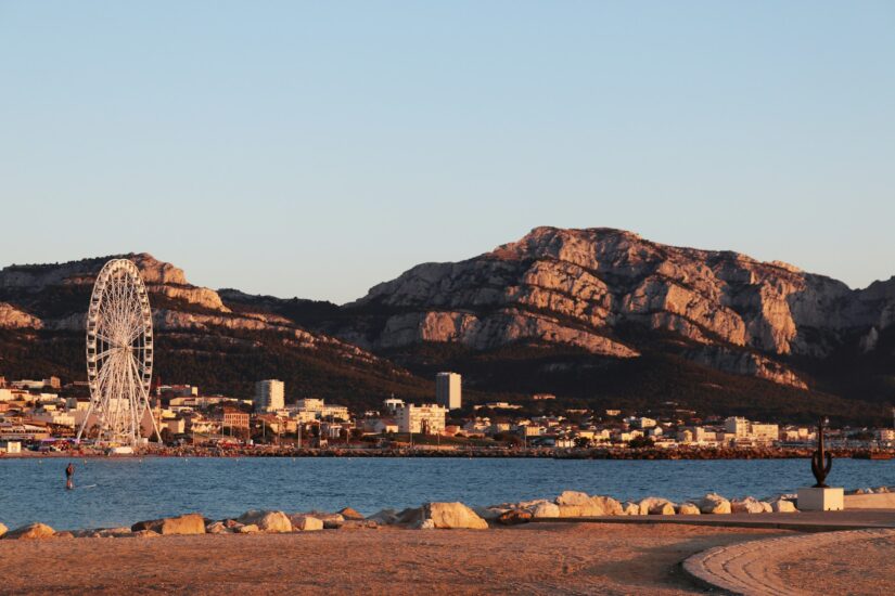 hotéis na Avenida du Prado e Castellane em Marseille na França Uma roda-gigante à beira-mar ao pôr do sol, evocando onde ficar em Marselha ao longo da Avenida du Prado ou no bairro Castellane, com prédios da cidade e montanhas atrás.
