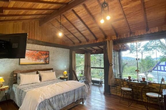 Quarto aconchegante em cabine de madeira com uma cama grande, área de estar e janelas grandes com vista para a natureza demonstra onde ficar em Miguel Pereira.