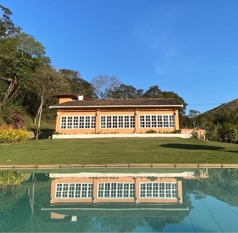 Uma casa com grandes janelas onde ficar em Miguel Pereira é refletida em uma piscina, cercada por grama e árvores sob um céu claro.