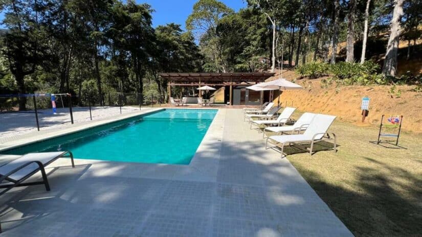 Piscina externa retangular com espreguiçadeiras, guarda-sóis e árvores ao fundo sob um céu claro.
