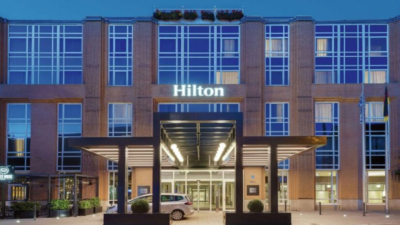 O exterior de um edifício do hotel Hilton onde ficar em Munique com janelas de vidro e uma entrada iluminada ao anoitecer.