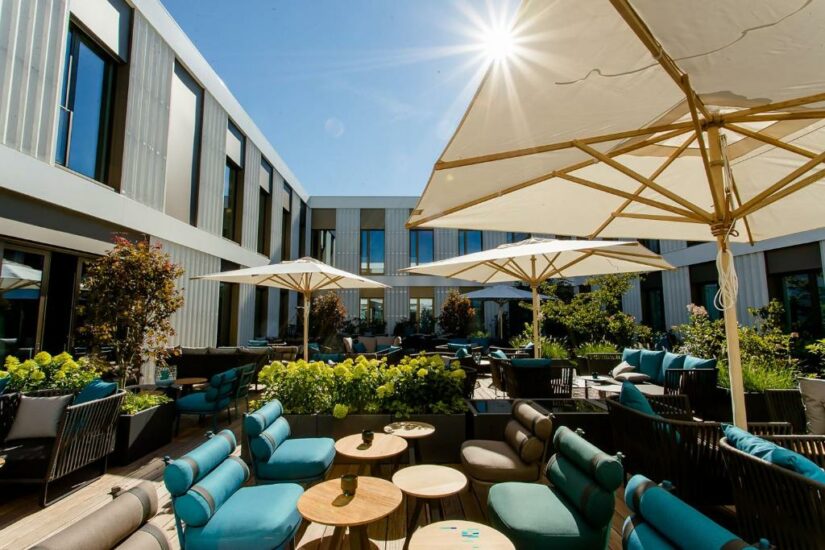 Área de lounge ao ar livre com assentos almofadados azuis, mesas, grandes guarda-chuvas e vegetação sob um céu ensolarado.