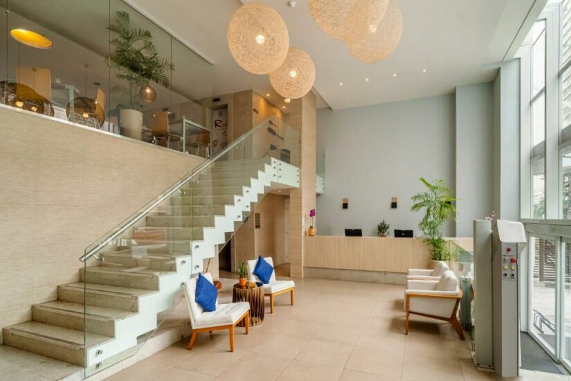 Saguão de hotel moderno Onde ficar em Niterói com escada de vidro, luzes penduradas, área de estar e balcão de recepção ao fundo.