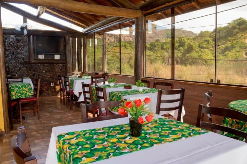 Pequeno restaurante com mesas de madeira, toalhas de mesa florais verdes e grandes janelas com vista para a vegetação e as colinas Onde ficar em Niterói