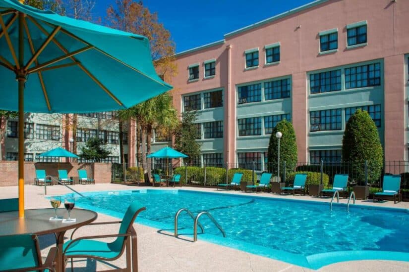 Piscina externa com espreguiçadeiras turquesa e guarda-sóis, ao lado de um moderno edifício rosa do hotel onde ficar em Nova Orleans em um dia ensolarado.
