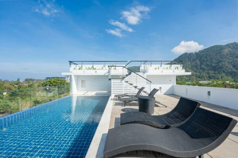Piscina infinita na cobertura com espreguiçadeiras pretas, guarda-corpo de vidro e vista para a montanha sob um céu azul.