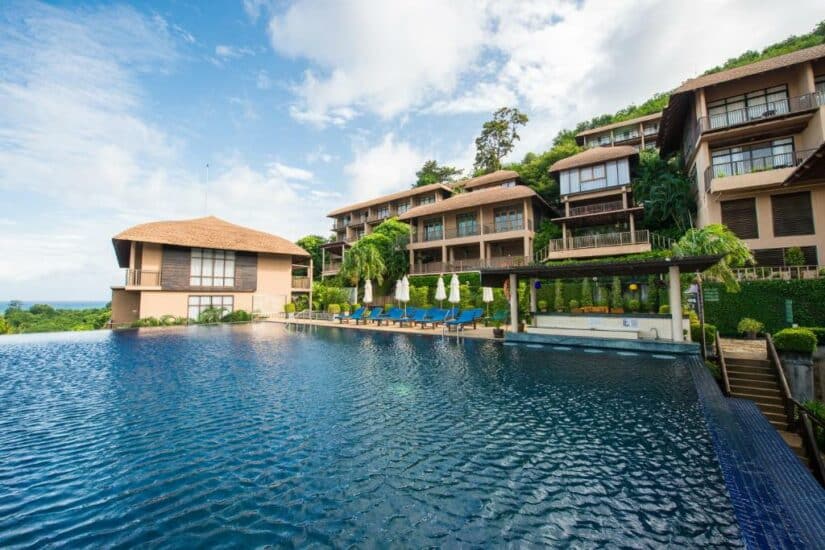 Grande hotel resort Onde ficar em Phuket com piscina de borda infinita, espreguiçadeiras e vegetação exuberante sob um céu parcialmente nublado.