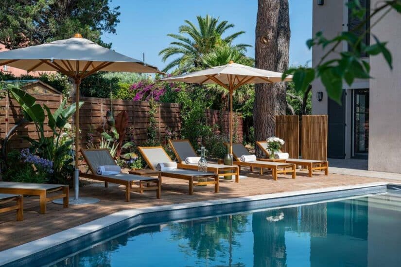 Espreguiçadeiras à beira da piscina com toalhas sob guarda-sóis, cercadas por vegetação e flores em um dia ensolarado demonstrando onde ficar em Saint Tropez no verão.