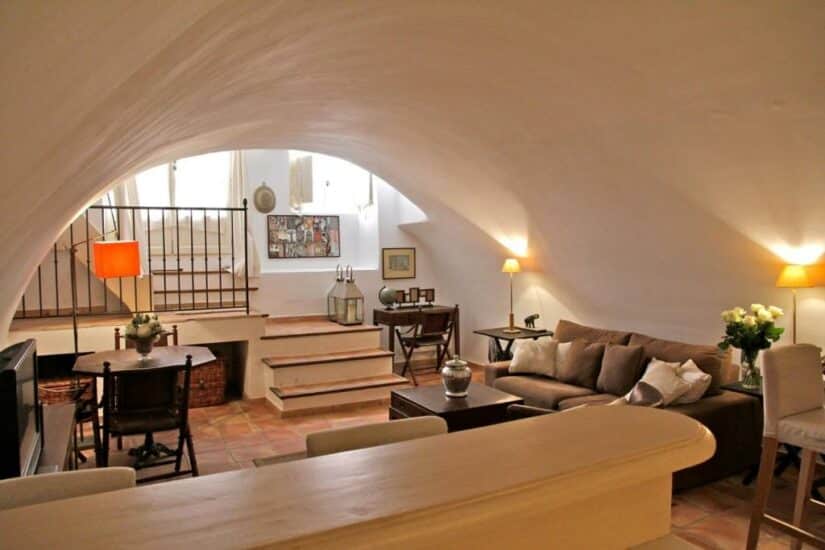 Sala de estar aconchegante de um dos hotéis da cidade histórica de Saint Tropez com teto em arco, iluminação aconchegante, móveis marrons e escadas que levam a uma área elevada.
