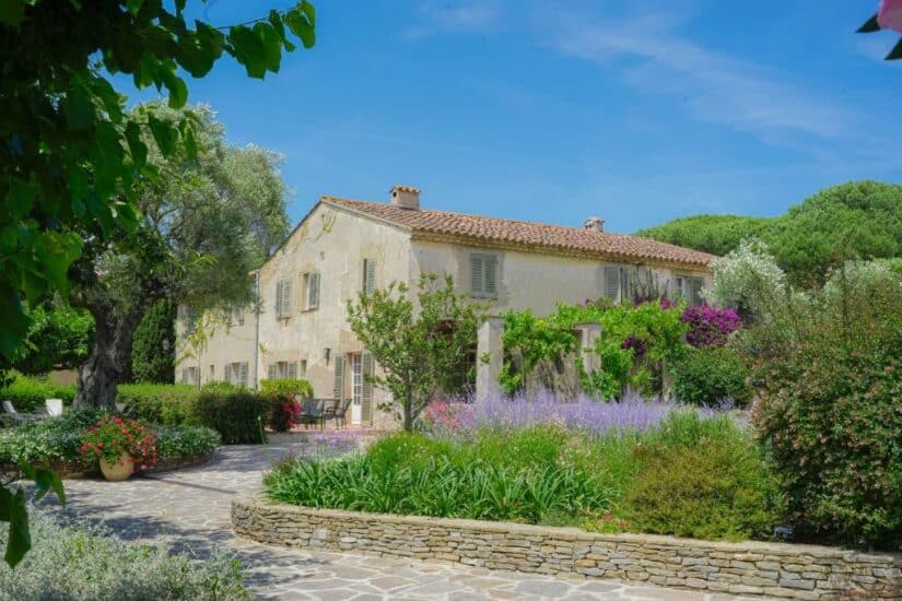 Uma casa de pedra onde ficar em Saint Tropez com telhado de telha cercada por jardins exuberantes e flores desabrochando sob um céu azul claro.