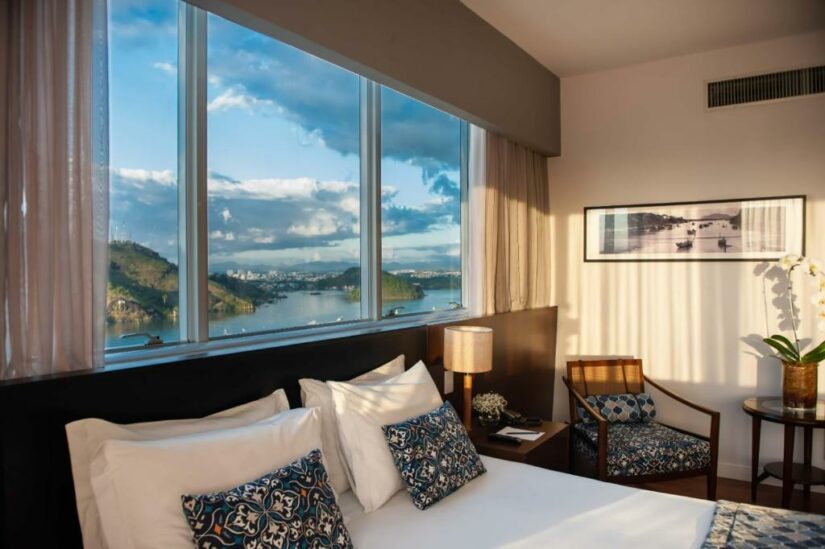 Um quarto de hotel onde ficar em Vitória aconchegante com uma grande janela que oferece uma vista panorâmica de um rio, colinas e céu azul.