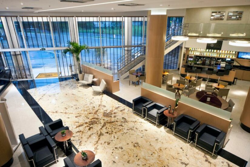 Lobby moderno do hotel com piso de mármore, assentos pretos, mesas pequenas e uma área de bar próxima a grandes janelas de vidro.
