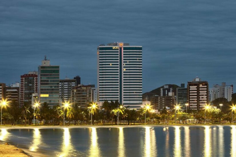 Paisagem urbana com edifícios altos  onde ficar em Vitória ao longo de uma orla à noite, palmeiras e luzes de rua refletindo na água.