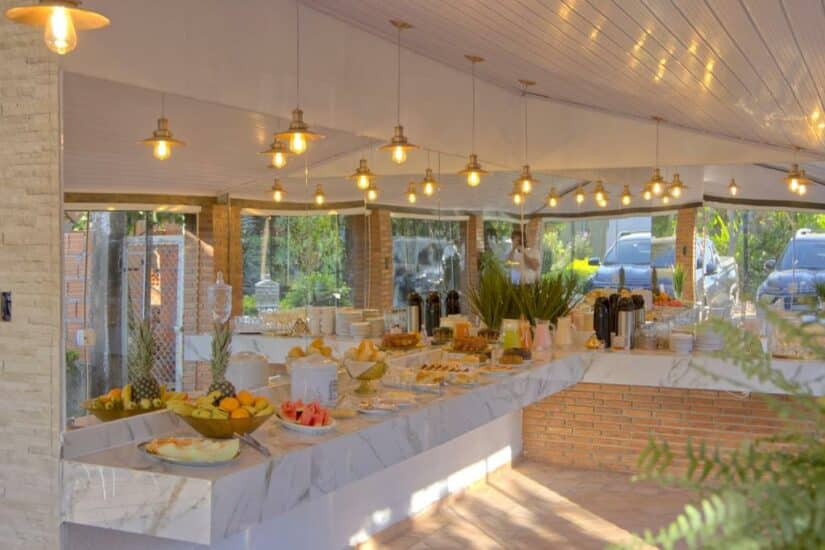 Uma mesa de bufê bem iluminada com frutas, doces e bebidas sob luzes penduradas em uma sala moderna com paredes de vidro onde ficar em Chapada dos Guimarães.