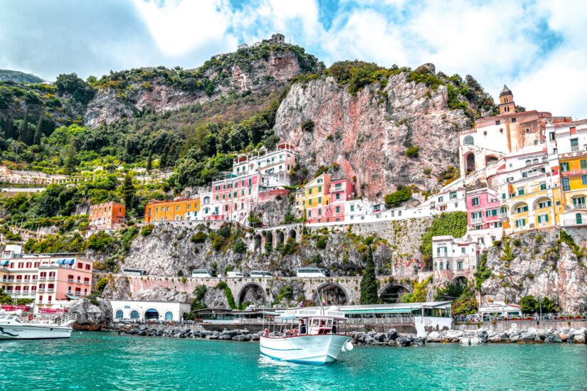 onde ficar na Costa Amalfitana em Amalfi Prédios coloridos em Amalfi em uma encosta íngreme acima da água azul-turquesa com barcos em primeiro plano.