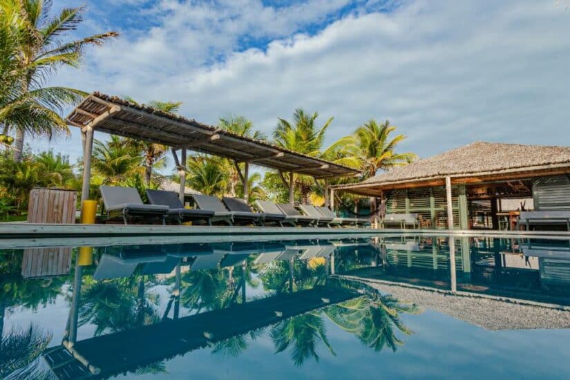 Espreguiçadeiras à beira de uma piscina com palmeiras e um prédio com telhado de palha sob um céu parcialmente nublado onde ficar em Canoa Quebrada.