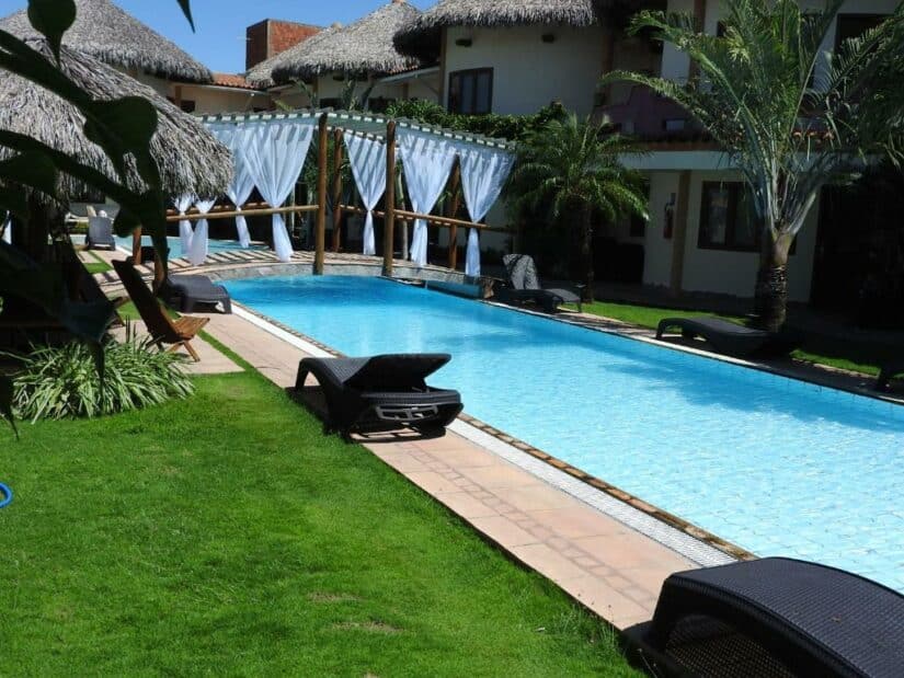 Uma piscina externa estreita com espreguiçadeiras, palmeiras e cortinas brancas ao lado de um prédio de resort tropical onde ficar em Canoa Quebrada.