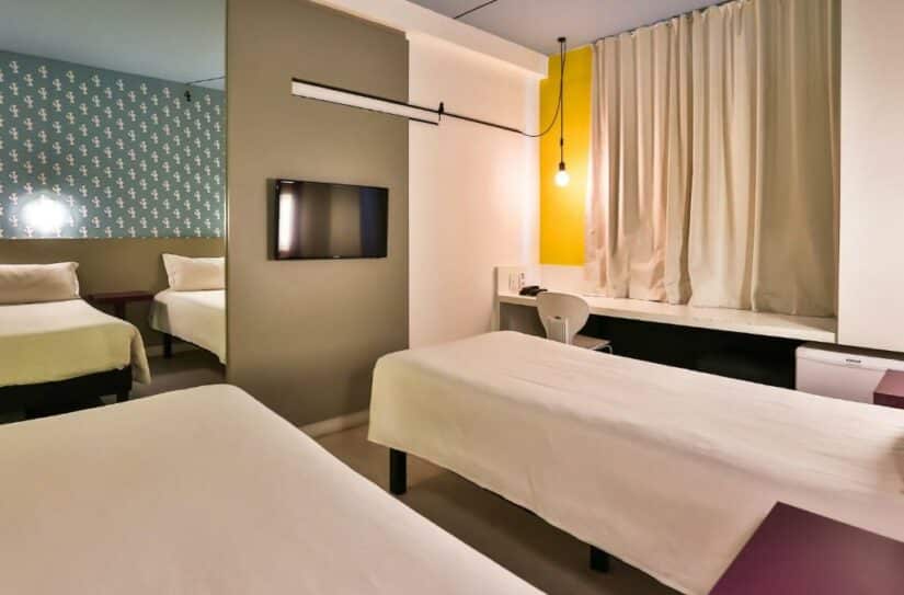 Quarto de hotel moderno com duas camas de solteiro, TV, escrivaninha, cadeira e janela grande com cortinas fechadas.