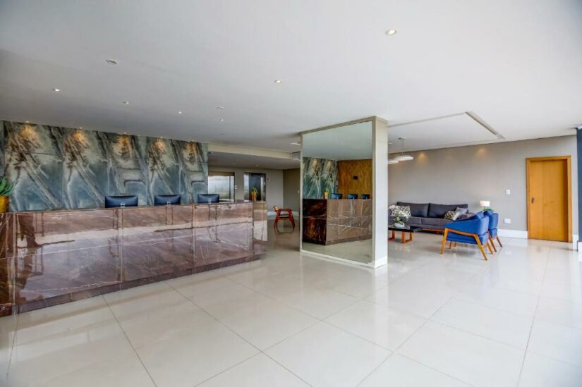Lobby moderno do hotel com balcão de recepção em mármore, espelho grande e uma área de estar com sofá azul.