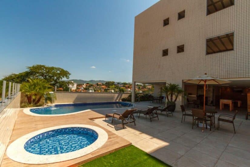 Área da piscina no terraço com espreguiçadeiras, banheira de hidromassagem e sala de jantar ao ar livre ao lado de um edifício moderno onde ficar em Inhotim.