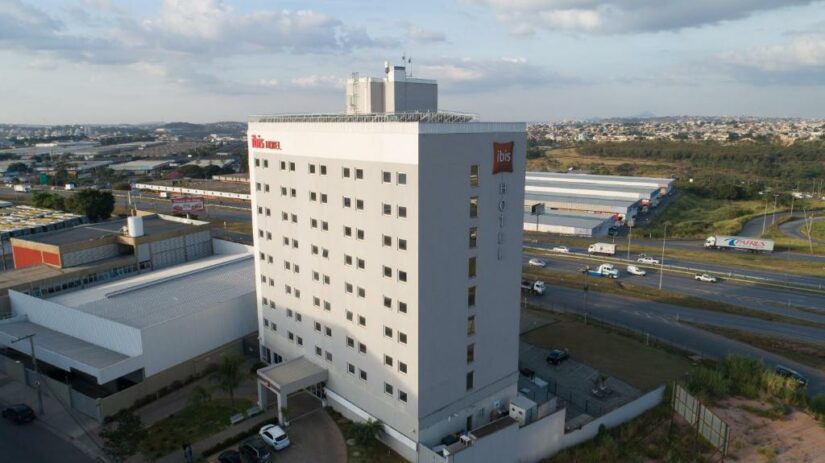 Vista aérea de um edifício alto e branco do hotel Ibis onde ficar em Inhotim próximo a uma rodovia movimentada com carros e vegetação ao redor.