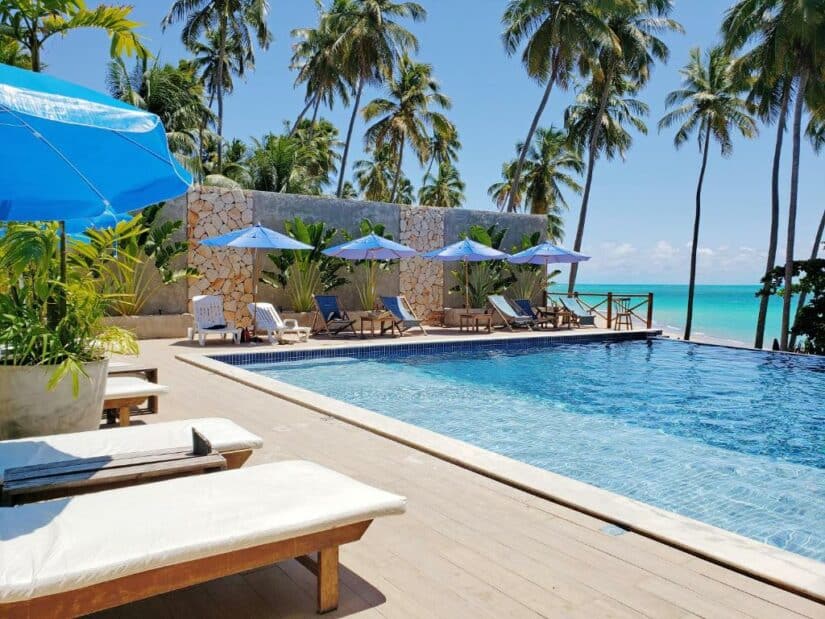 Espreguiçadeiras e guarda-sóis à beira da piscina em uma praia tropical onde ficar em Japaratinga com palmeiras e o oceano azul claro ao fundo.