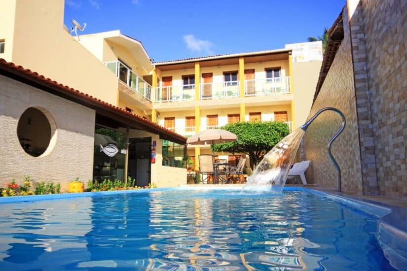 Piscina no pátio com cascata, cercada por um pátio, cadeiras e um prédio amarelo de dois andares onde ficar em Japaratinga.