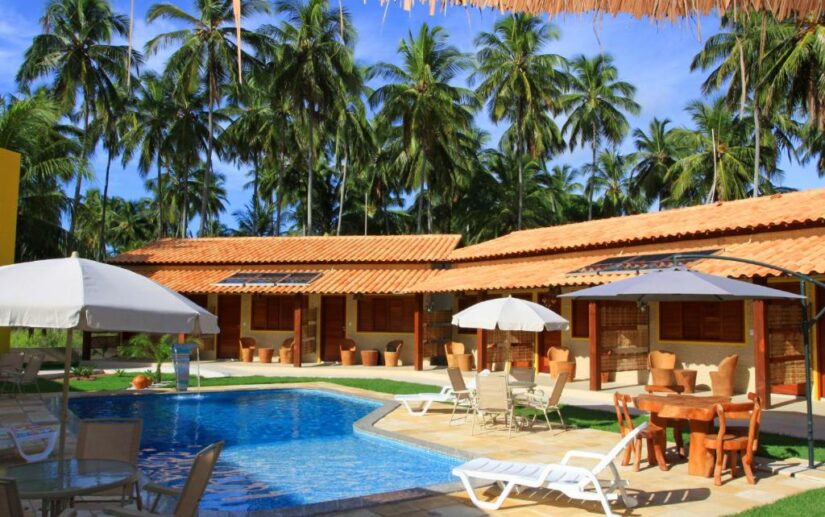 Piscina externa com espreguiçadeiras e guarda-sóis, cercada por palmeiras e edifícios com telhado de terracota.