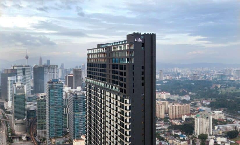 Edifício alto e preto do hotel Aila onde ficar em Kuala Lumpur com arranha-céus da cidade e um céu nublado ao fundo.