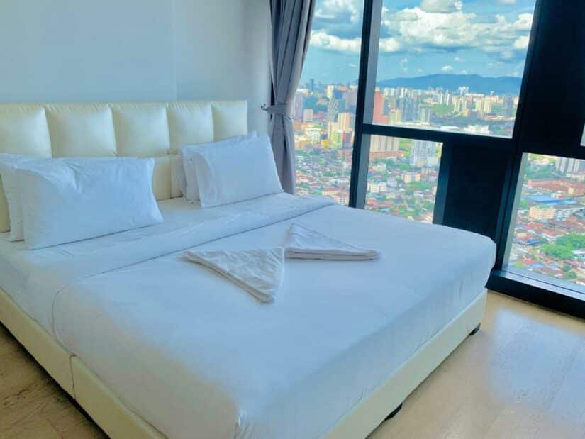 Uma cama bem arrumada em um quarto bem iluminado com grandes janelas com vista para o horizonte da cidade onde ficar em Kuala Lumpur em um dia ensolarado.