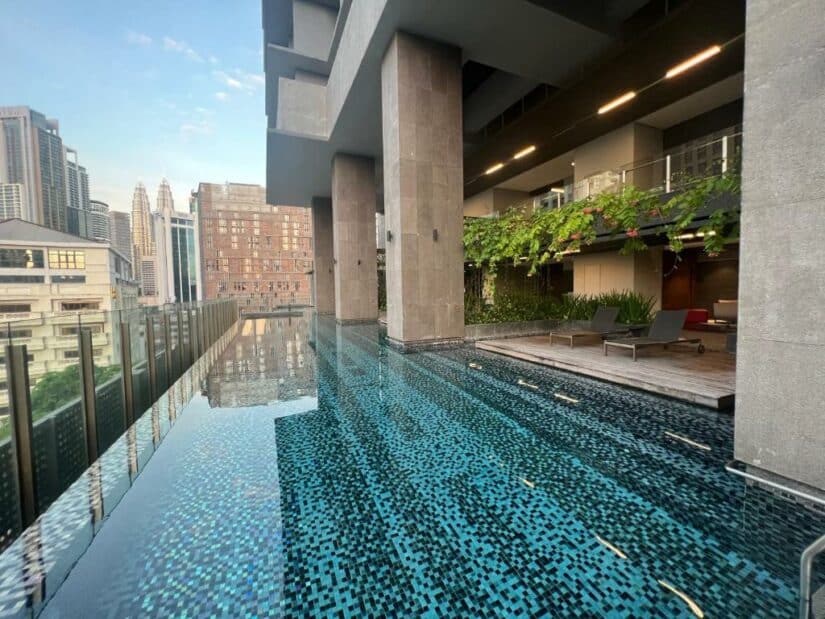 Piscina de borda infinita com mosaicos ao lado de um edifício moderno, com os arranha-céus da cidade visíveis ao fundo.