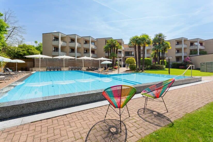 Piscina externa com espreguiçadeiras, guarda-sóis, palmeiras e cadeiras coloridas em um pátio ensolarado do hotel.