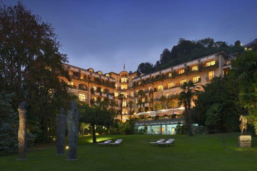 Um grande hotel onde ficar em Lugano com janelas brilhantes ao entardecer, cercado por árvores e cadeiras de gramado em um jardim gramado.
