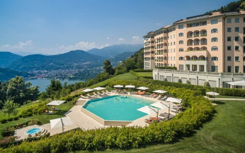 Hotel de luxo onde ficar em Lugano em uma colina com piscina, espreguiçadeiras, lago e montanhas ao fundo sob um céu claro.