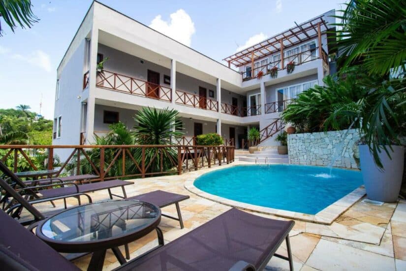 Pátio moderno do hotel com piscina, espreguiçadeiras, plantas tropicais e um prédio branco de dois andares.