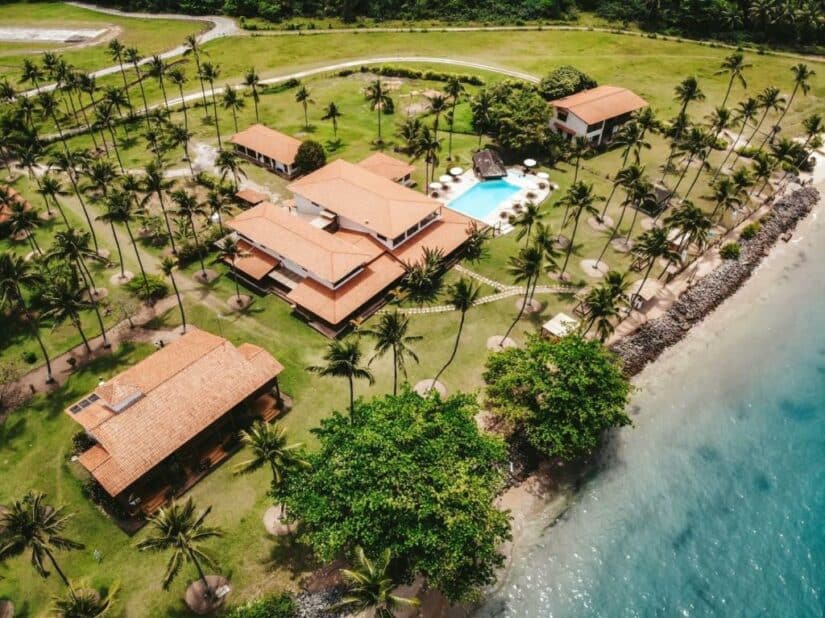 Vista aérea de um resort à beira-mar onde ficar em Morro de São Paulo com edifícios de telhado vermelho, palmeiras e uma piscina à beira-mar.