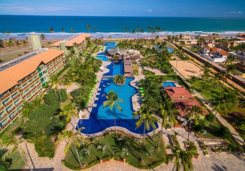 Vista aérea de um resort tropical Onde ficar em Muro Alto com uma grande piscina, palmeiras e praia ao longo do oceano.
