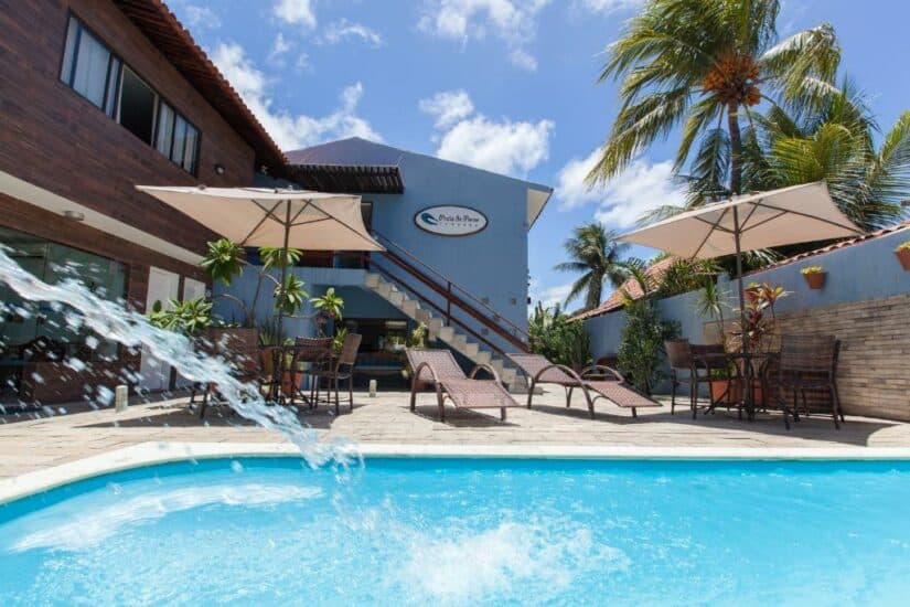 Piscina ensolarada com espreguiçadeiras, guarda-sóis, plantas tropicais e um prédio azul ao fundo.
