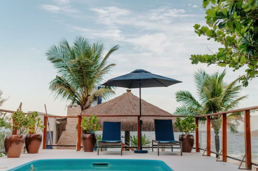 Duas espreguiçadeiras e um guarda-sol ao lado de uma piscina com palmeiras e um telhado de palha à beira-mar.