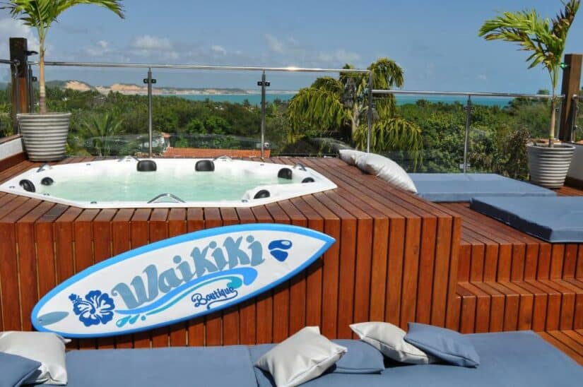 Jacuzzi ao ar livre em um deck de madeira com almofadas azuis, palmeiras e uma prancha de surfe lendo Waikiki onde ficar em Pipa.