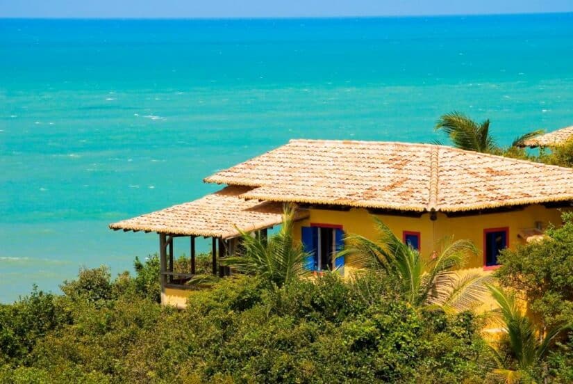 Uma casa amarela com telhado de telha fica em meio à vegetação, com vista para a água azul-turquesa do oceano sob um céu claro.