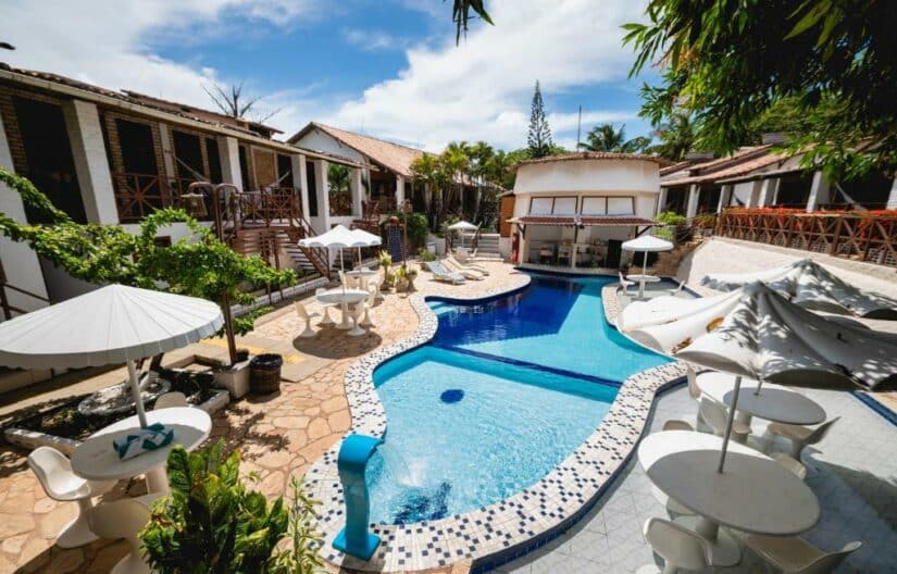 Piscina curvilínea com guarda-sóis brancos e espreguiçadeiras em um resort onde ficar em Pipa cercado por vegetação tropical e edifícios.