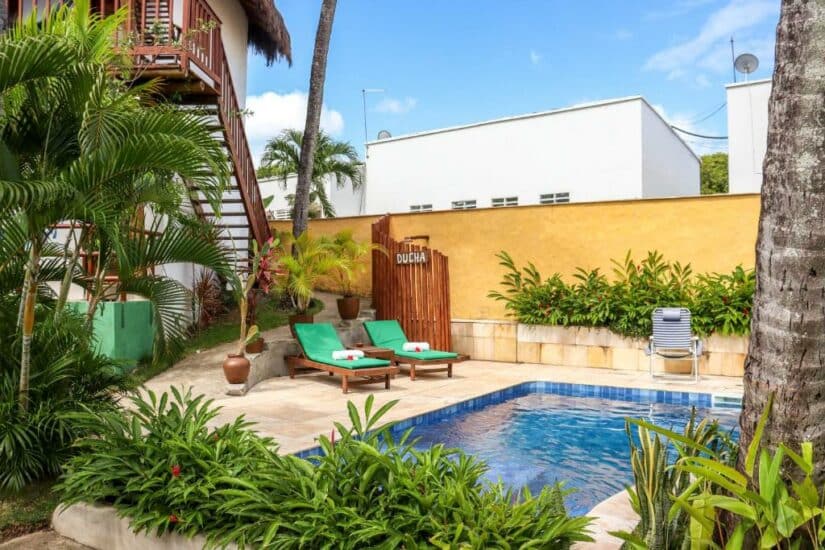 Uma pequena piscina externa com duas espreguiçadeiras verdes, cercada por plantas tropicais e uma ducha de madeira.