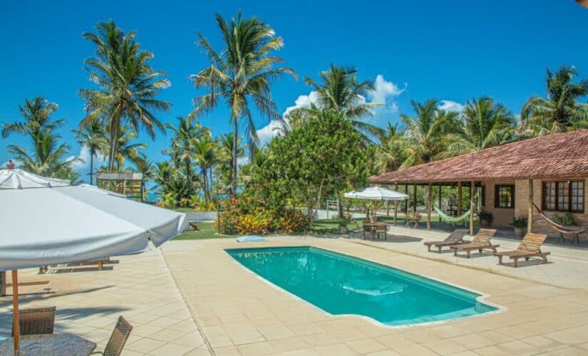 Uma cena tropical à beira da piscina com espreguiçadeiras, guarda-sóis, palmeiras e um prédio com redes sob um céu ensolarado.