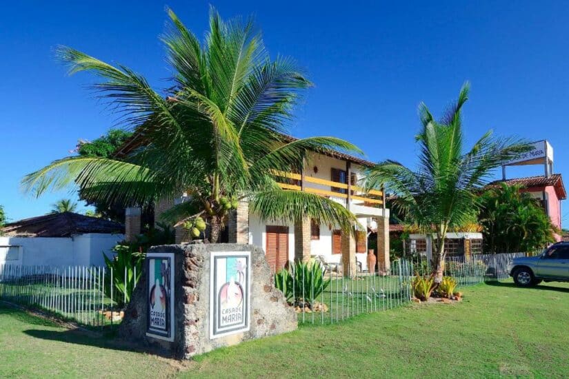 Pousada tropical onde ficar em Prado com palmeiras, uma varanda amarela e um pátio gramado sob um céu azul claro.