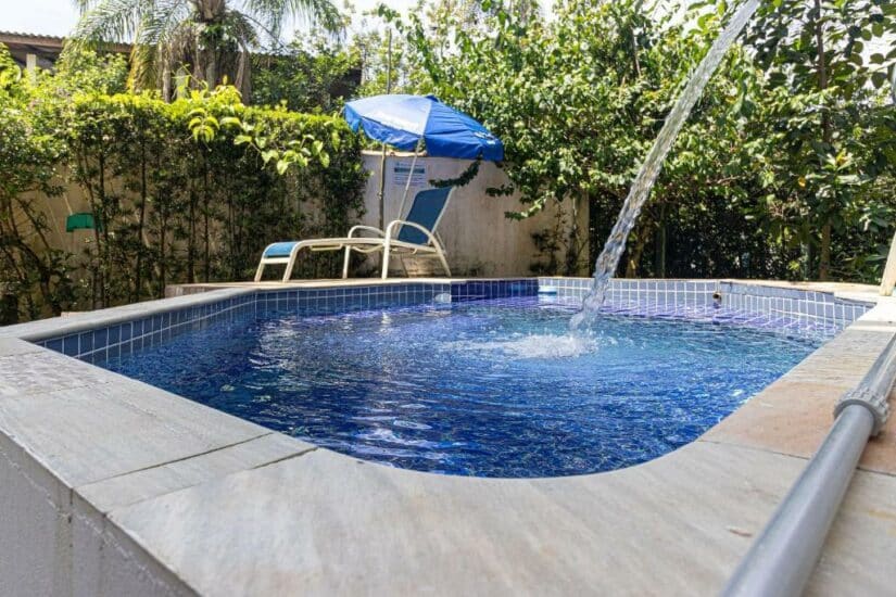 Pequena piscina no quintal com água limpa, espreguiçadeiras e um guarda-sol azul cercado por plantas verdes.