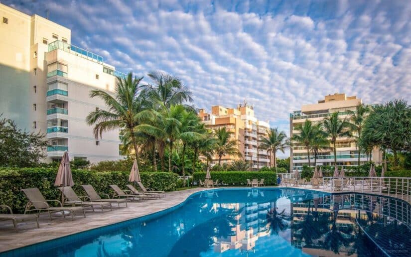 Piscina externa com espreguiçadeiras, palmeiras e edifícios modernos onde ficar em Riviera de São Lourenço sob um céu com nuvens dispersas.