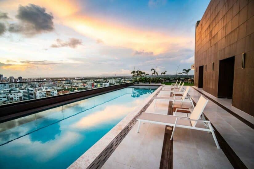 Piscina na cobertura onde ficar em Santo Domingo com espreguiçadeiras com vista para o horizonte da cidade ao pôr do sol, com nuvens refletindo na água.
